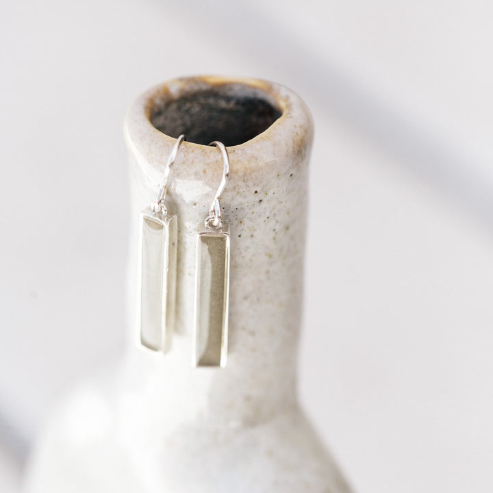 a pair of memorial bar earrings with ashes in sterling silver hanging from the lip of a thin speckled vase