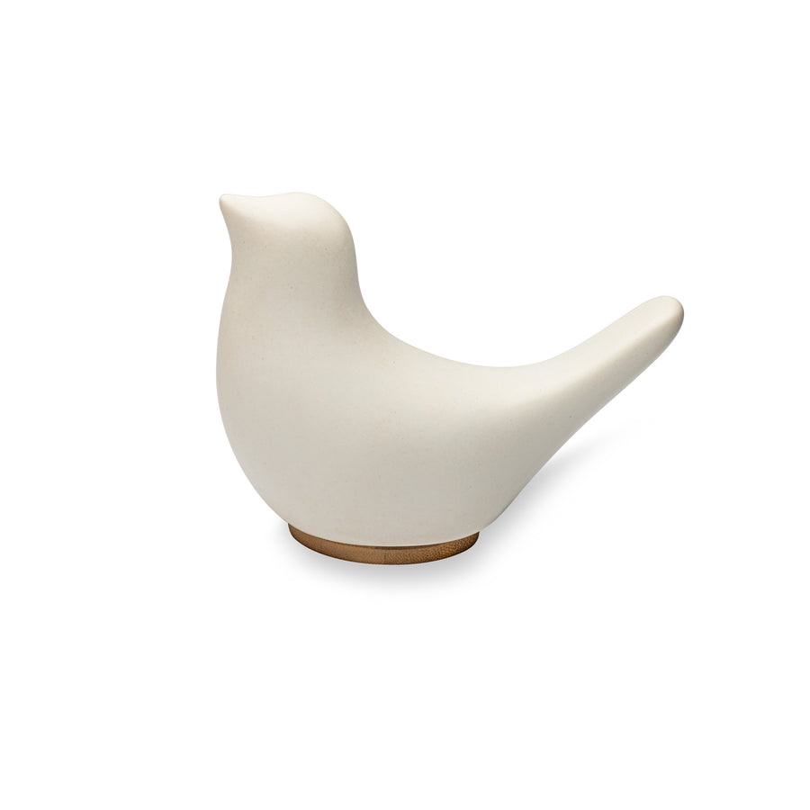 A side view of Close By Me's Ceramic Bird Urn facing to the left, against a solid white background. The urn has a matte white finish and its bamboo lid is visible at the bottom, doubling as a small pedestal.