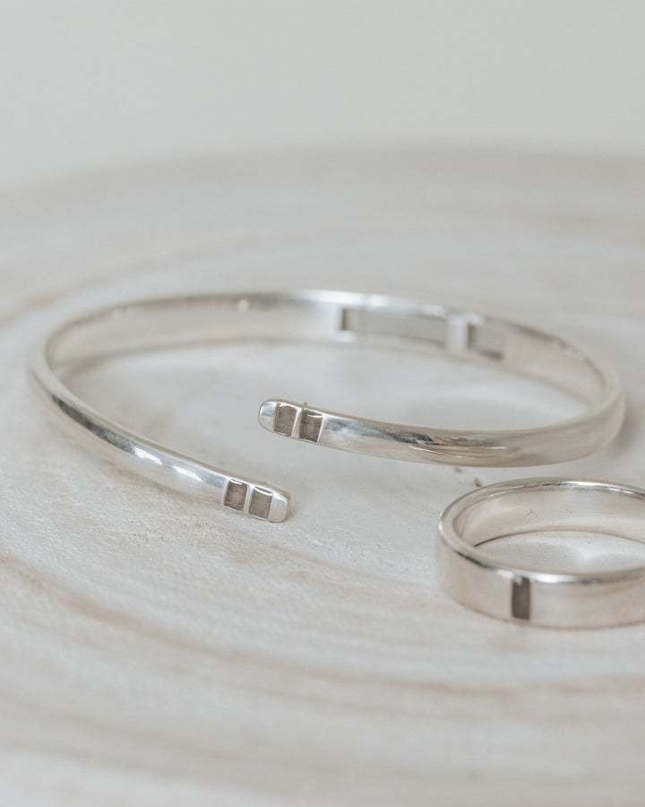 A photo showing the bypass hinged cuff ashes bracelet and the simple band cremation ring in sterling silver lying next to each other against a gray background