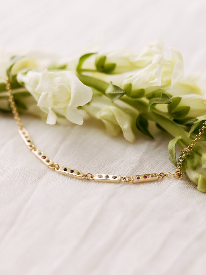 The Tribute Bracelet by Close By Me in 14K Yellow Gold on a light mesh fabric background. It has 5 bar links with 3 birthstones in each.