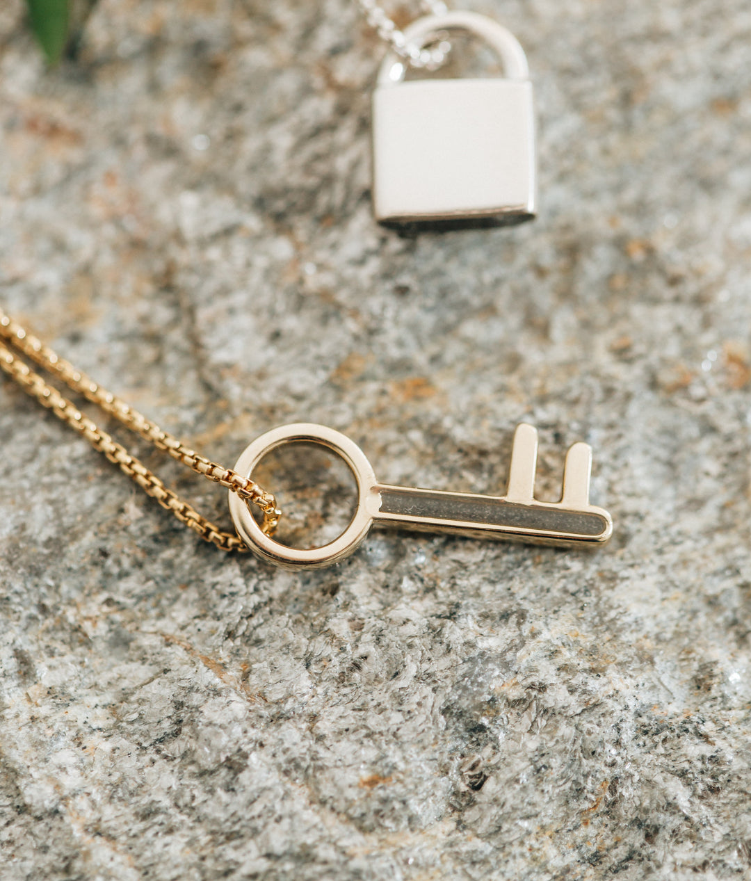 Pictured here is close by me jewelry's Key Cremains Pendant design in 14K Yellow Gold lying flat on a multi-colored textured surface next to the Sterling Silver Lock Cremation Pendant