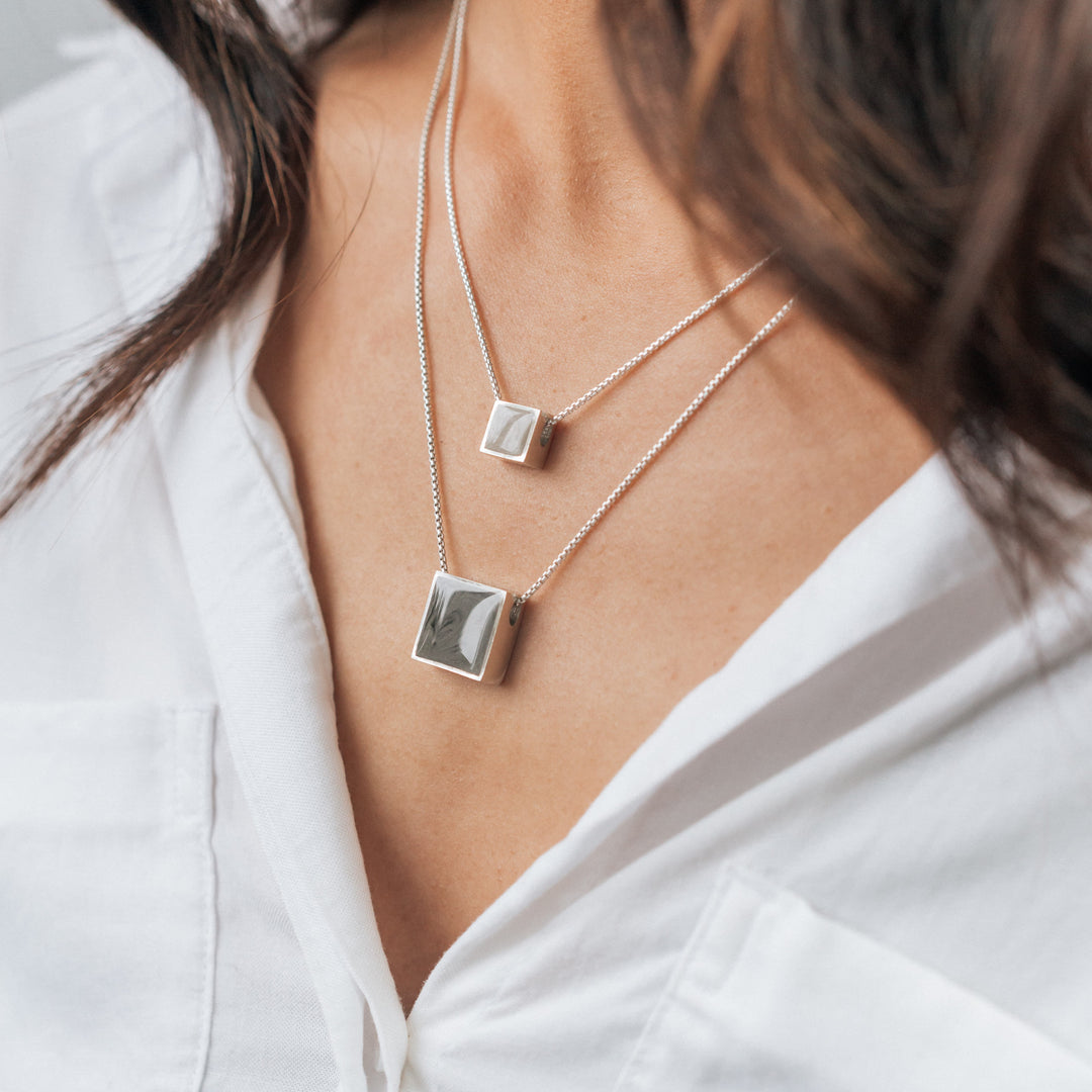 A model with tan skin in a white blouse wearing close by me's Sterling Silver Sliding Cremains Pendants, one with a Small Square Setting and one with a Large Square Setting