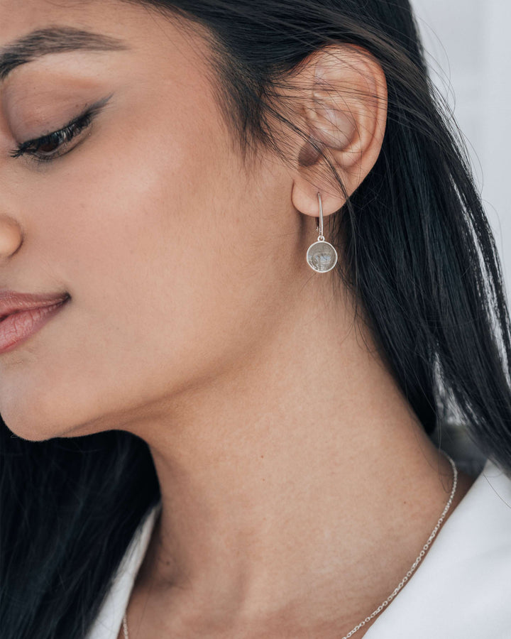 A photo of a woman with medium-brown skin tone and dark looking to the side and down while wearing sterling silver circular dangle cremation earrings