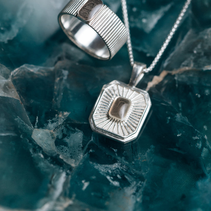 A close-up, overhead view of Close By Me's Tessa Cremation Necklace and matching ring set on a dark green agate-style backdrop.