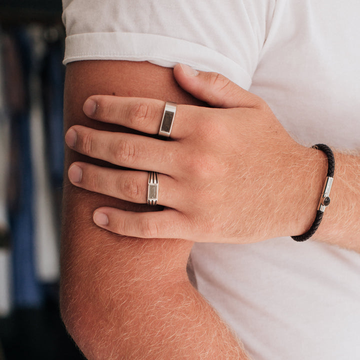 male model wearing close by me's classic band cremation ring on index finger, ridged band rectangle signet cremation ring on ring finger, and leather cord cremation bracelet