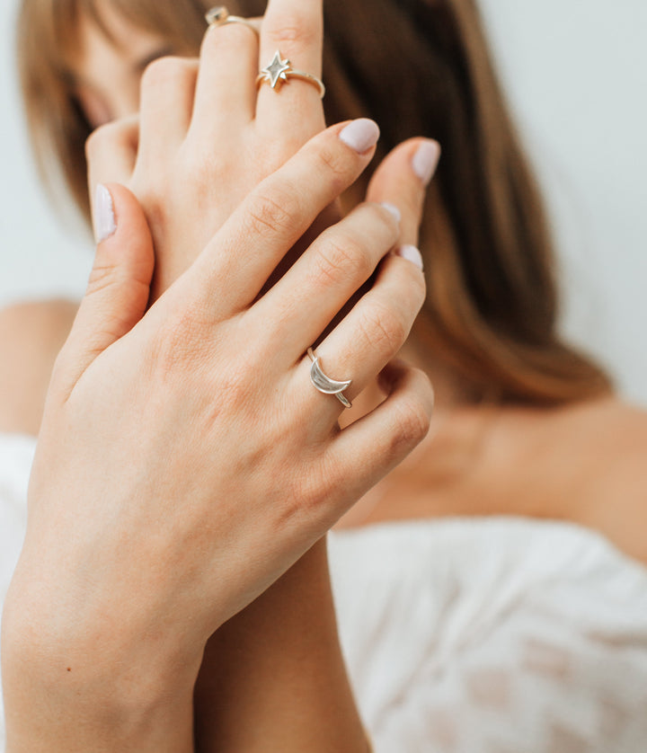 This photo shows a light-skinned model wearing several pieces of cremation jewelry in mixed metals by close by me jewelry. Her hands are held out in front of her to block the view of her face and the background is blurred. Her right hand is held on top of the left and shows the Crescent Moon Ashes Ring in Sterling Silver with a light gray cremation setting. Her left hand wears both the 14K Yellow Gold North Star Ring and a Circular Stacking Ring