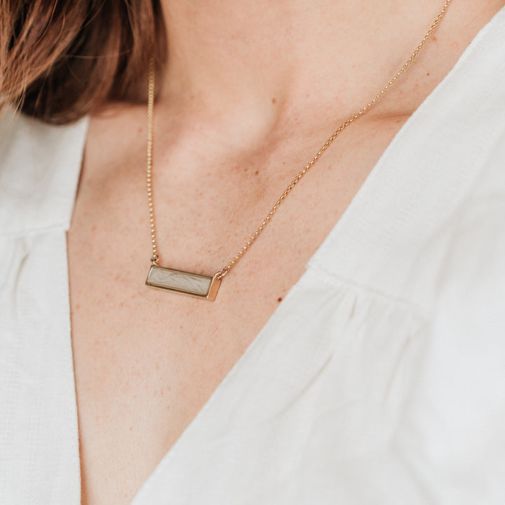Pictured here is a model in a white blouse wearing the 14K Yellow Gold Lateral Bar Ashes Pendant designed by close by me jewelry