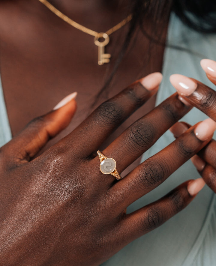 This photo shows a model with dark skin and hair in a blue v-neck blouse wearing close by me jewelry's 14K Rose Gold and Champagne Diamond Oval Split Shank Cremation Ring design.  Blurred in the background, you can see the Key Ashes Pendant design around her neck..