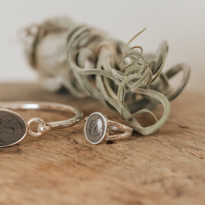 This is a stylized photo that shows the Sterling Silver Oval Split Shank cremation Ring design by close by me jewelry on a wooden table in front of an air plant