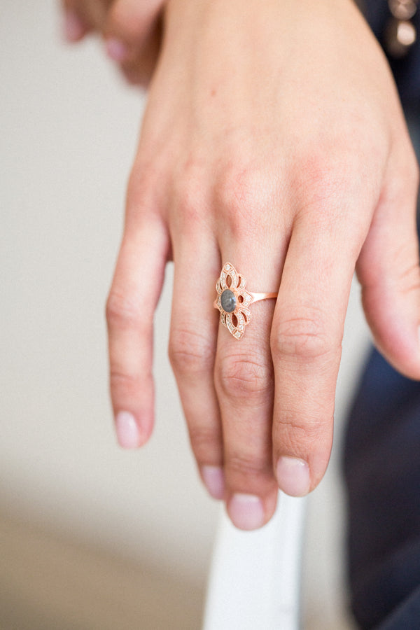 A close-up of Close By Me's vintage-style World War II Cremation Ring in 14K Rose Gold with Champagne Diamonds being worn on the middle finger of a female model's hand. The oval bezel in the center contains solidified cremated remains that are gray in color with a subtle swirled pattern visible.