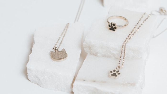 From left to right: Close By Me's Cat Cremation Necklace, Paw Print Stacking Cremation Ring, and Small Paw Print Cremation Necklace resting atop glistening white chunks of stone.