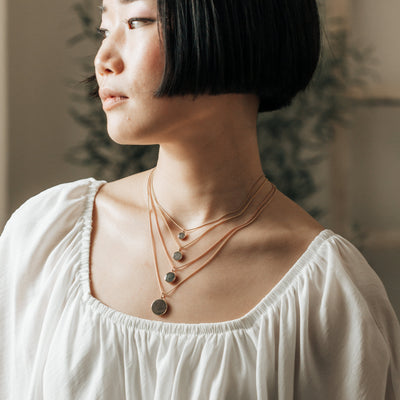 A model gazes to the side, wearing Close By Me's 6mm, 8mm, 10mm, and 15mm Dome Cremation Pendants in 14k gold. Each pendant is worn on a different length chain so that they are layered from smallest to largest.