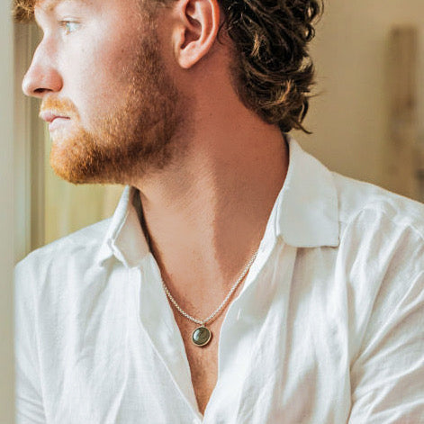 A male model in a white button down shirt looking out of a window to his right as he wears a sterling silver cremation necklace made with ashes by close by me