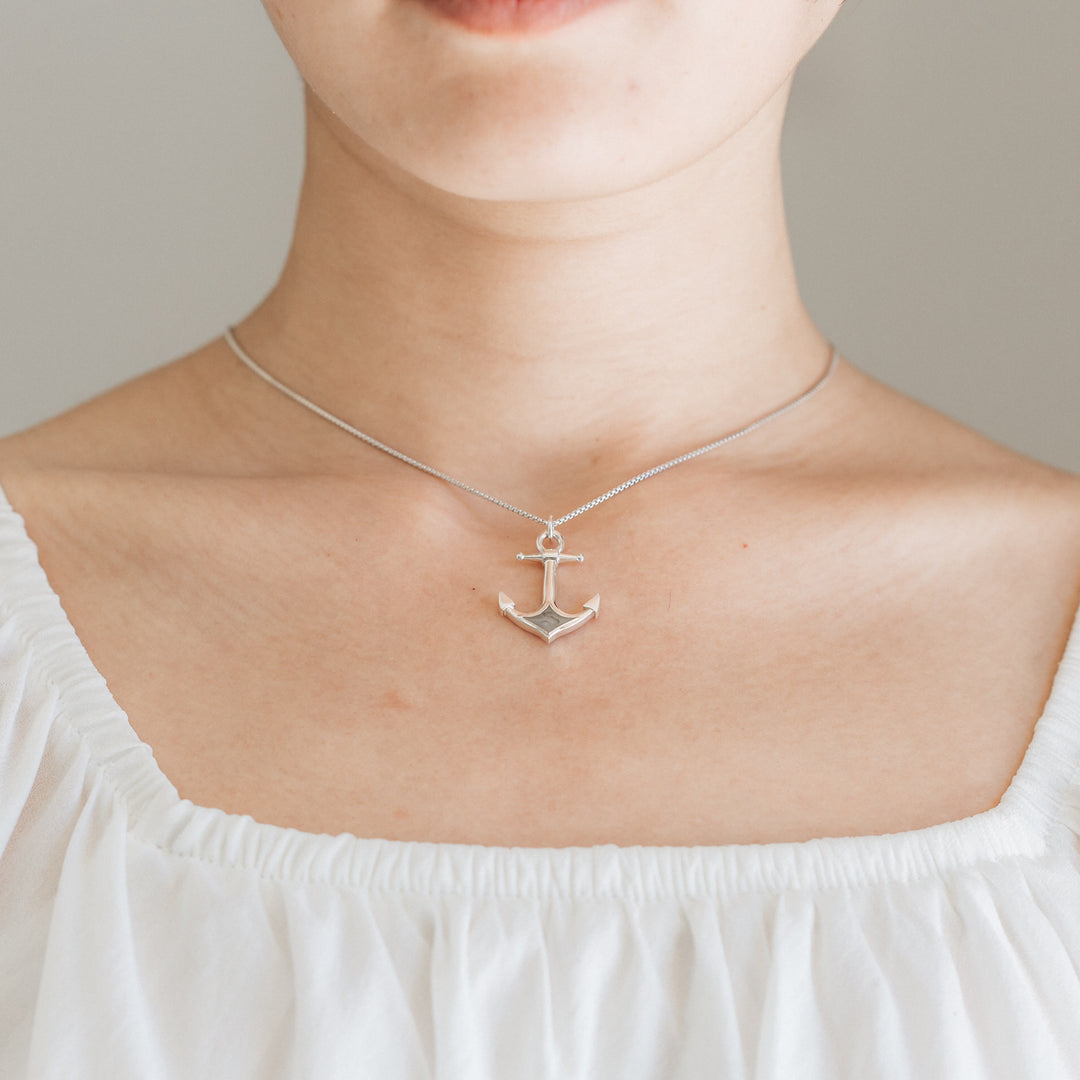 Hanging on a thin chain, Close By Me's Anchor Cremation Necklace rests against the collarbone of a light-skinned woman shown from her chin to the top of her white blouse.