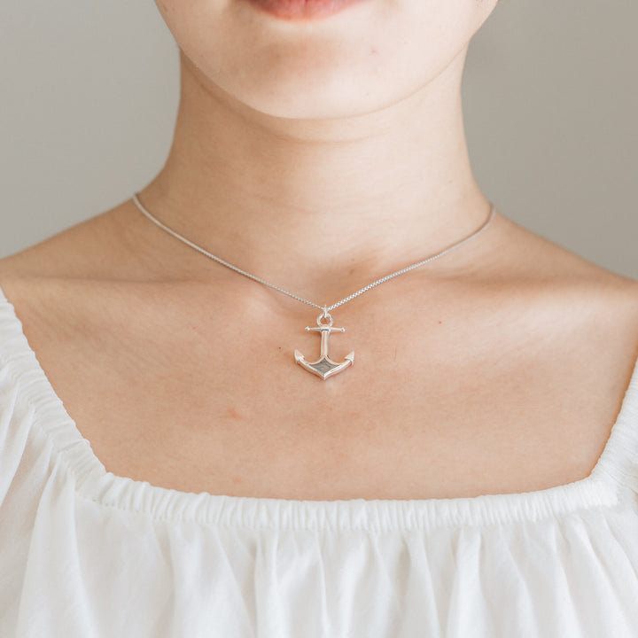 Hanging on a thin chain, Close By Me's Anchor Cremation Necklace rests against the collarbone of a light-skinned woman shown from her chin to the top of her white blouse.