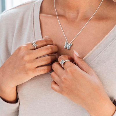 This photo shows a warm skinned model in a light heather top wearing rings and pendants with cremated remains in Sterling Silver by close by me jewelry; around her neck is the Butterfly Pendant