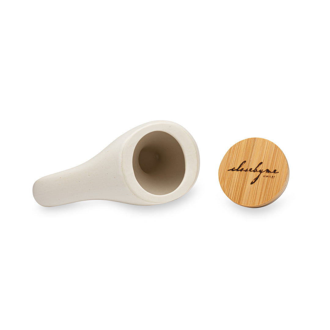 Close By Me's Ceramic Bird Urn lays on its side, with its bottom facing the camera, giving an inside view of the urn. Its Close By Me Jewelry-branded bamboo lid rests upright beside it.