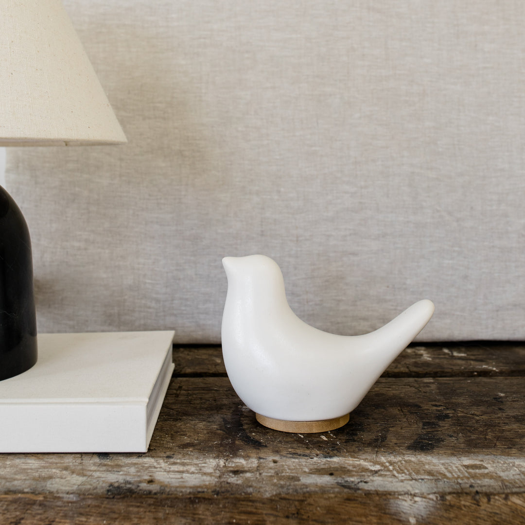 White ceramic bird cremation urn on a wooden surface next to a lamp on top of a book