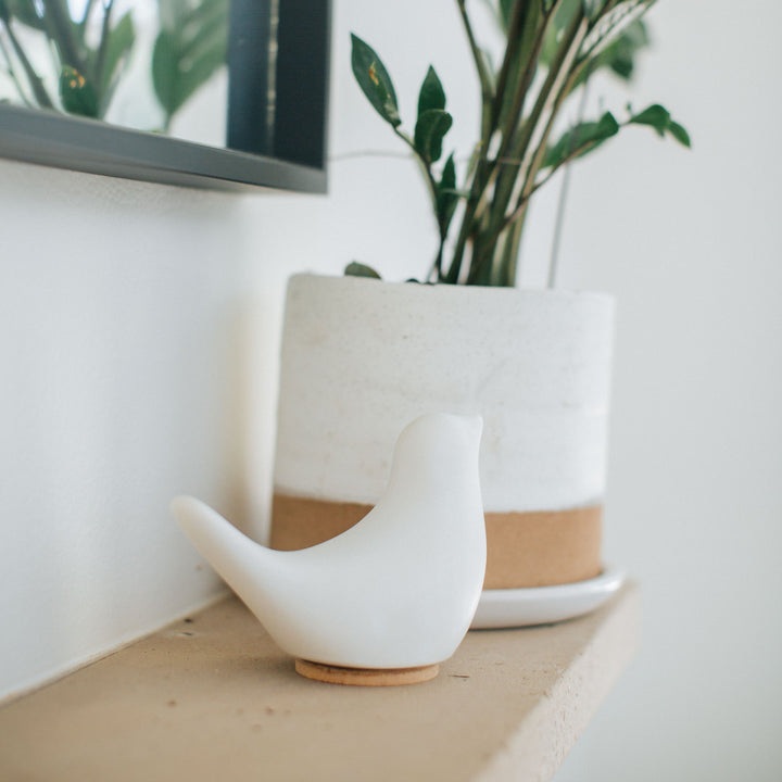 White ceramic bird cremation urn on a mantel, underneath a mirror and next to a potted plant
