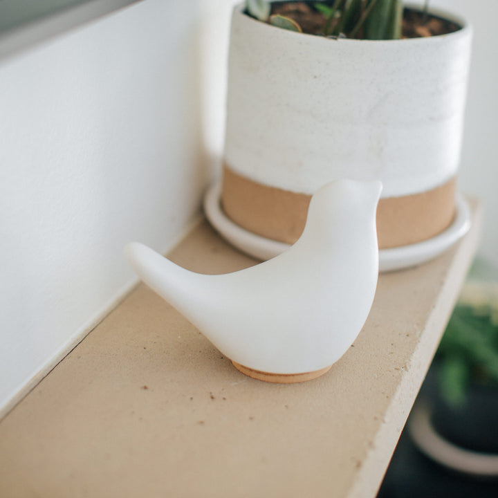 White ceramic bird cremation urn on a mantel next to a potted plant