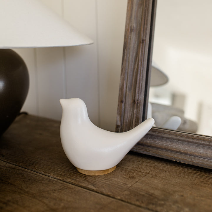 White ceramic bird cremation urn on a wooden surface with a mirror and lamp in the background