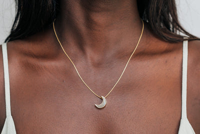 A  close-up of Close By Me's Crescent Moon Cremation Necklace in 14K Yellow Gold around the neck of a dark-skinned model.