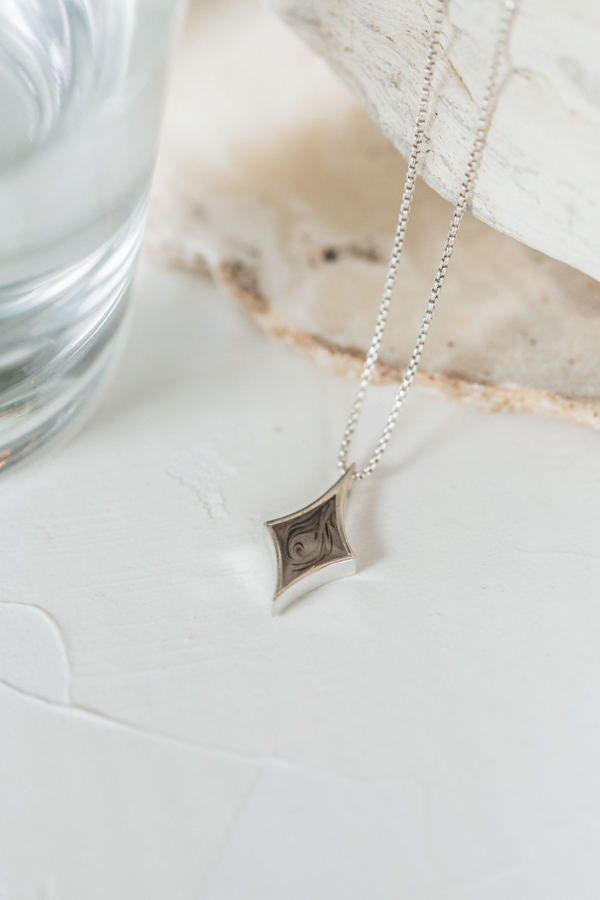 This photo shows the Sterling Silver Diamond Cremains Pendant laying flat on a white multi-textured background