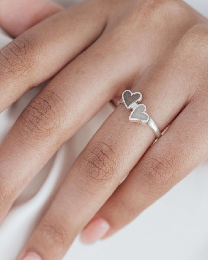 A cremation ring with double heart setting designs, set with ashes, being worn on a model's ring finger in sterling silver