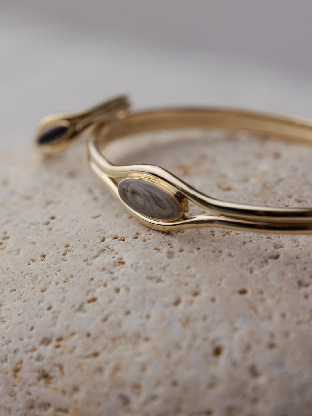 Close-up of the Double Band Oval Bangle Cremation Bracelet on a beige stone with the Double Band Oval Cremation Ring propped up against it, out of focus.