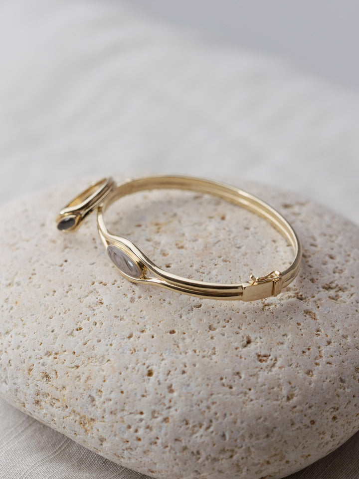 The Double Band Oval Bangle Cremation Bracelet on a beige stone with the Double Band Oval Cremation Ring propped up against it.