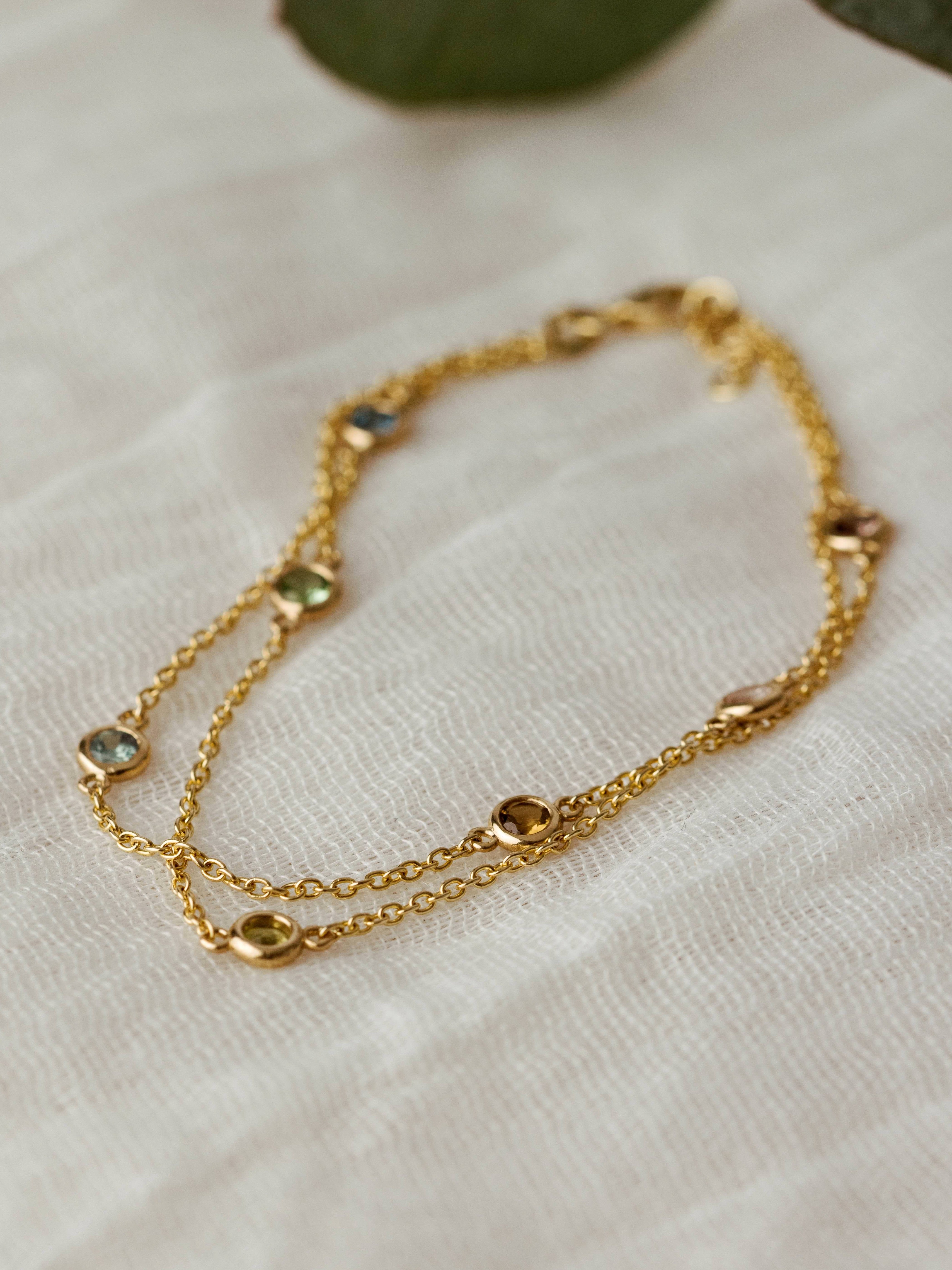 Close By Me's Double Strand Birthstone Bracelet in 14k yellow gold set with 7 different stones laying on light mesh fabric.