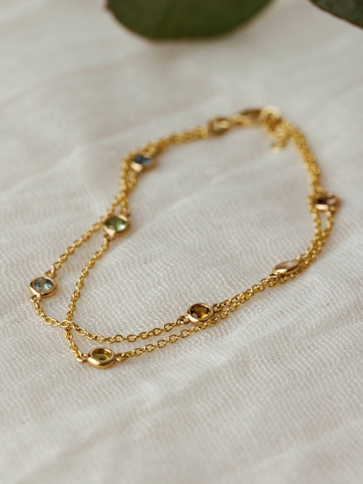 Close By Me's Double Strand Birthstone Bracelet in 14k yellow gold set with 7 different stones laying on light mesh fabric.