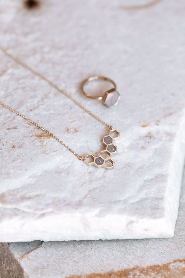 This photo shows the Hexagon Cluster Necklace with ashes by close by me jewelry lying next to the Large Hexagon Ashes Stacking Ring design on a flat piece of white marble; both pieces have been cast in 14K Yellow Gold and set with cremated remains