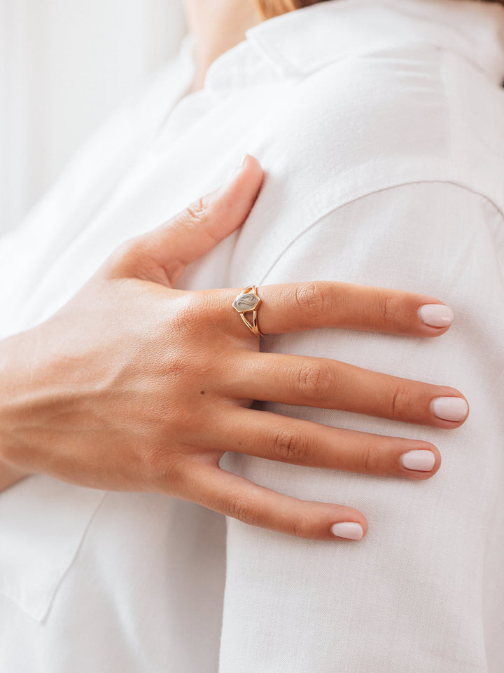 The hand of a young, olive skinned woman in a white blouse rests against her shoulder, Close By Me's Hexagon Split Shank Cremation Ring on her index finger.