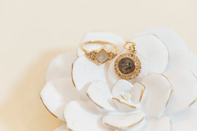 Close By Me's Kite Setting Diamond Cluster Cremation Ring and Large Diamond Halo Cremation Necklace, both in 14K Yellow Gold with Champagne Diamonds, resting atop a white ceramic flower.