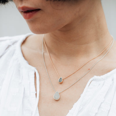 A light skinned model with dark hair wearing both the Small Pear Sliding Cremated Remains Necklace in 14K Rose Gold and the Large Pear Sliding Memorial Pendant in Sterling Silver by close by me jewelry