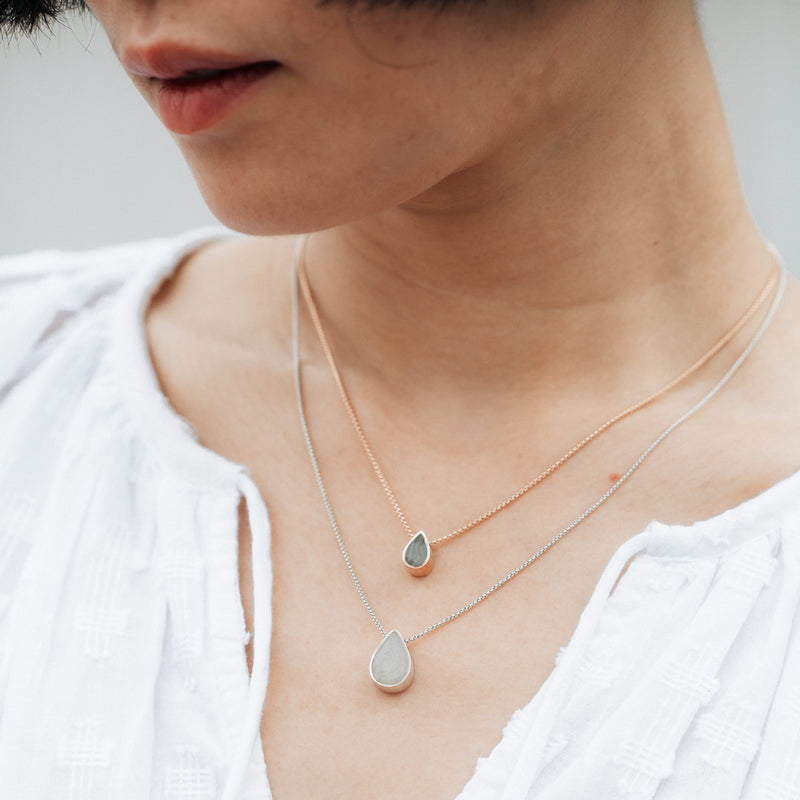 A light skinned model with dark hair wearing both the Small Pear Sliding Ashes Necklace in 14K Rose Gold and the Large Pear Sliding Cremains Necklace in Sterling Silver