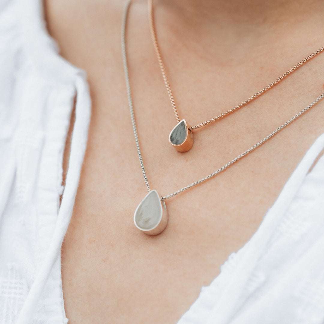 A close up of the Small Pear Sliding Ashes Necklace in 14K Rose Gold and the Large Pear Sliding Cremains Necklace in Sterling Silver by close by me being worn by a light skinned model in white top