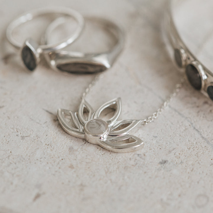 This photo shows several Sterling Silver pieces of ashes jewelry by close by me jewelry lying flat on a porous gray stone; the Lotus Flower Cremains Necklace design is featured in the center