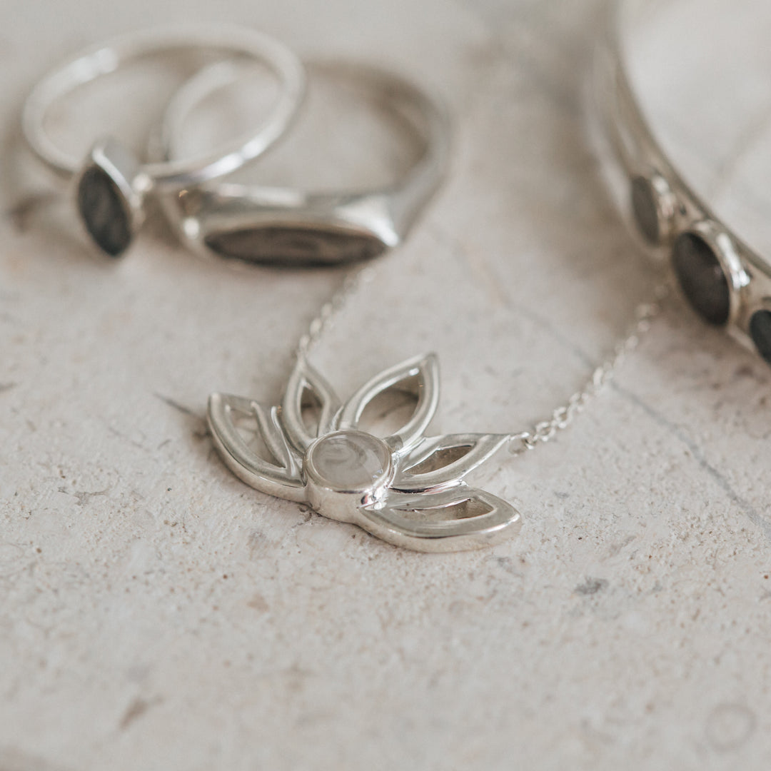 This photo shows several Sterling Silver pieces of jewelry with ashes by close by me jewelry lying flat on a porous gray stone; the Lotus Flower Cremated Remains Necklace design is featured in the center