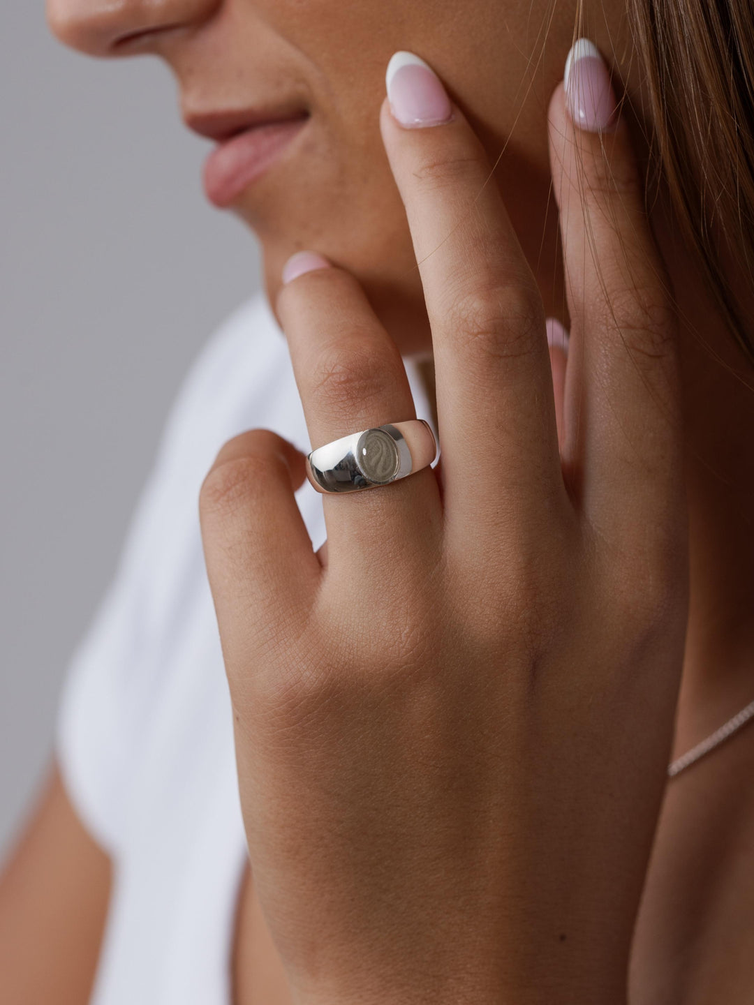 A woman is resting her hand against her face, wearing the Modern Oval Signet Cremation Ring on her ring finger.