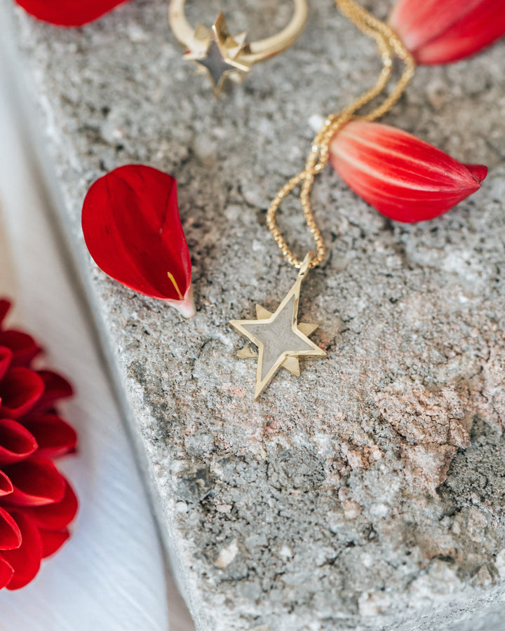 The North Star Pendant with ashes designed in 14K Yellow Gold by close by me jewelry lying flat on a gray textured surface with red flower petals