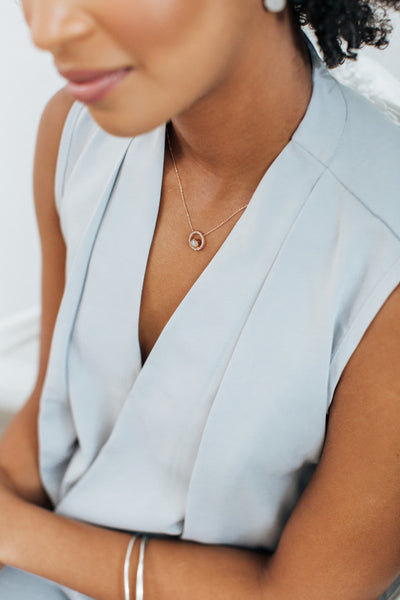 Overhead view of a dark-skinned, female model in a low-cut, V-neck, light blue-grey blouse wearing Close By Me's Open Diamond Halo Cremation Necklace around her neck. 