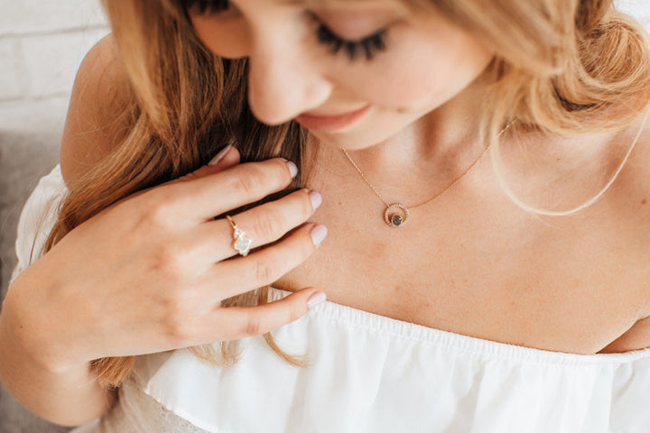An overhead view of a light-skinned, female model in an off-the-shoulder white blouse, wearing Close By Me's Open Diamond Halo Cremation Necklace around her neck. Her hand is resting against her chest and visible on her middle finger is Close By Me's Kite Setting Diamond Cluster Cremation Ring.