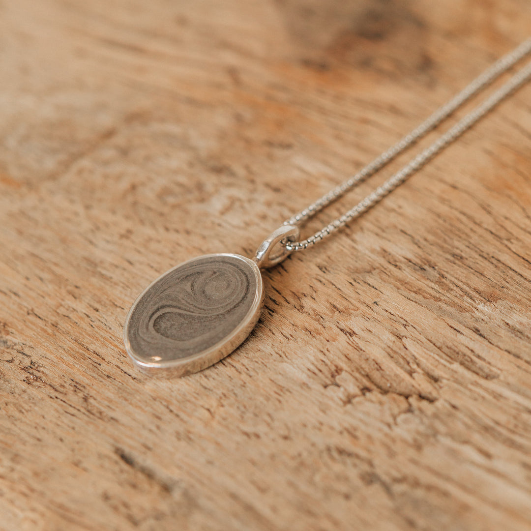The simple bail oval cremation necklace in sterling silver by close by me jewelry lying flat on a wooden surface 