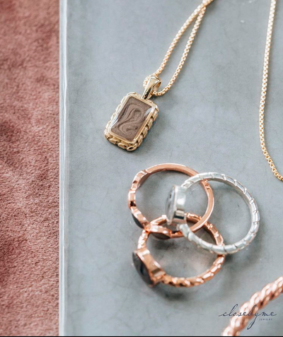 A close up of several pieces of close by me's cremation jewelry lying flat on a blue ceramic plaque; the focus is the Small Cable Cremation Necklace in 14K Yellow Gold