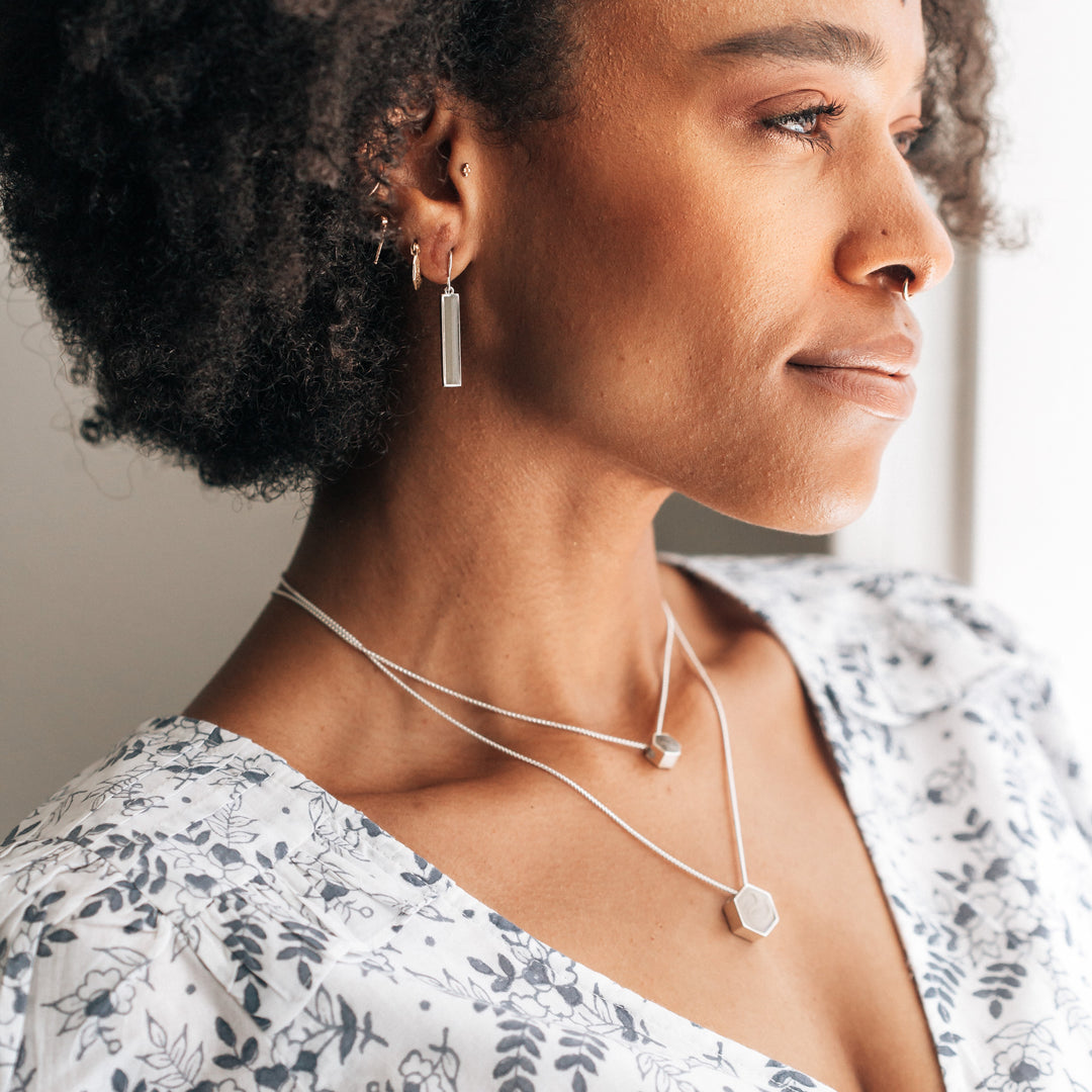 Pictured here are both the Large and Small Sterling Silver Hexagon Sliding Necklaces with cremains being worn by a dark skinned model in a white dress with gray flower patterns. She also wears several other pieces of jewelry with ashes designed by close by me jewelry in this photo.