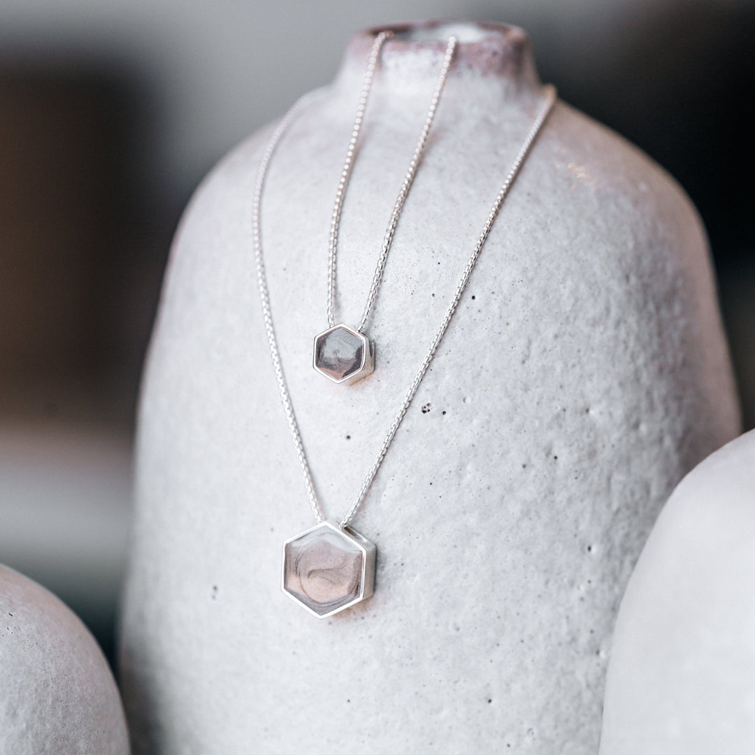 This photo shows the Sterling Silver Hexagon Sliding Pendant with Cremated Remains in both large and small sizes, designed by close by me jewelry, hung on a rough-cast white vase
