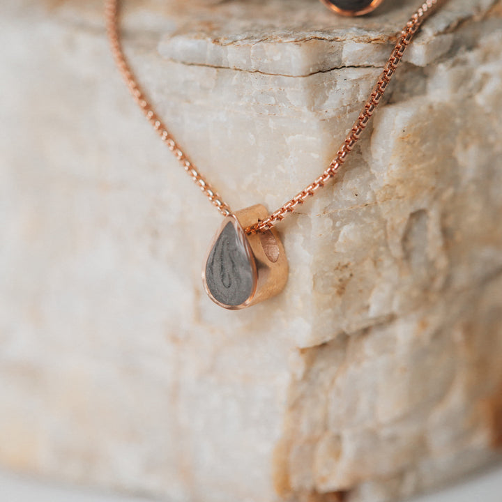 This photo shows a close up of close by me jewelry's Small Pear Sliding Ashes Pendant design in 14K Rose Gold hanging from a light warm-toned flaky stone