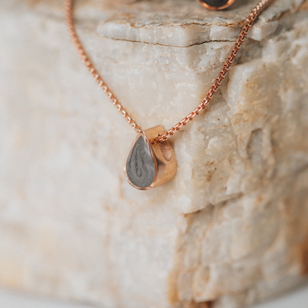 This photo shows a close up of close by me jewelry's Small Pear Sliding Cremated Remains Pendant design in 14K Rose Gold hanging from a light warm-toned flaky stone